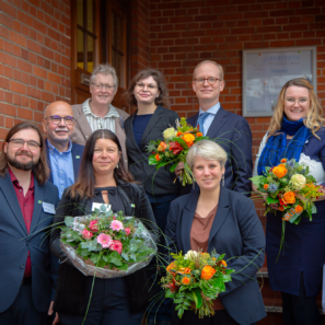 Eine Gruppe von Menschen feiert die Eröffnung der Pflegeschule in Hassel. Sie stehen aufgereiht auf der Eingangs-Treppe mit dem roten Backsteingebäude hinter ihnen. Vier von Acht Personen halten Blumensträuße in den Händen. Alle Lächeln geradewegs in die Kamera.